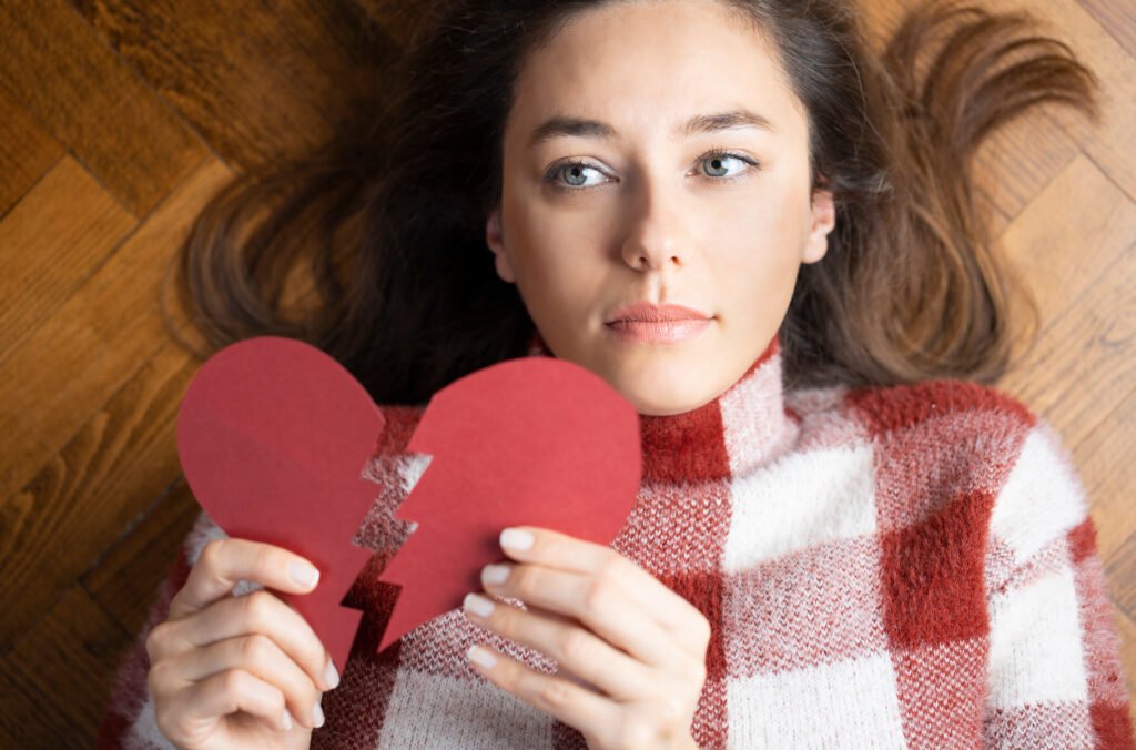 Woman lying on the floor holding a broken paper heart, looking sad and reflective after a breakup.