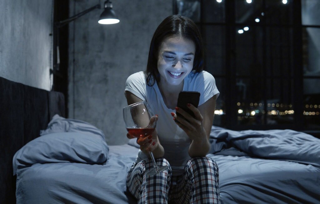 Woman sitting on her bed at night, smiling at her phone while holding a glass of wine, engaged in an online dating conversation.