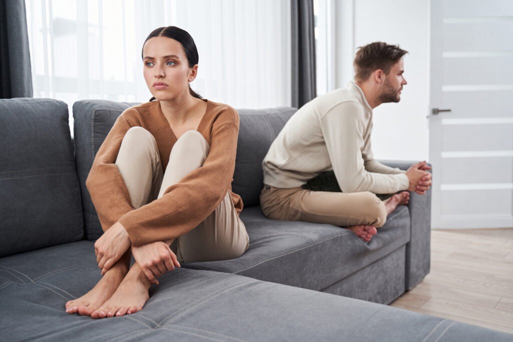 Man and woman sitting apart on a couch facing away from each other, showing emotional distance and tension in their relationship, as though one feels emotionally manipulated.