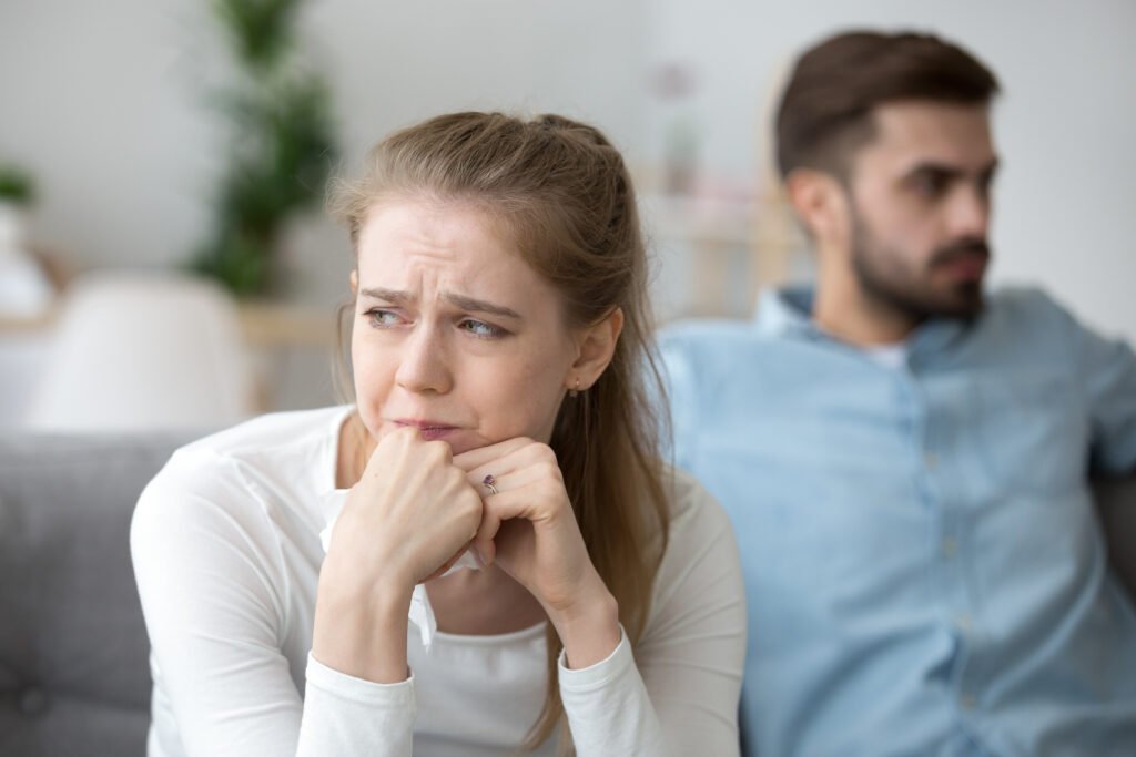 Woman trying to recover from a break up, sitting on a couch with a tissue while her partner avoids eye contact in the background.
