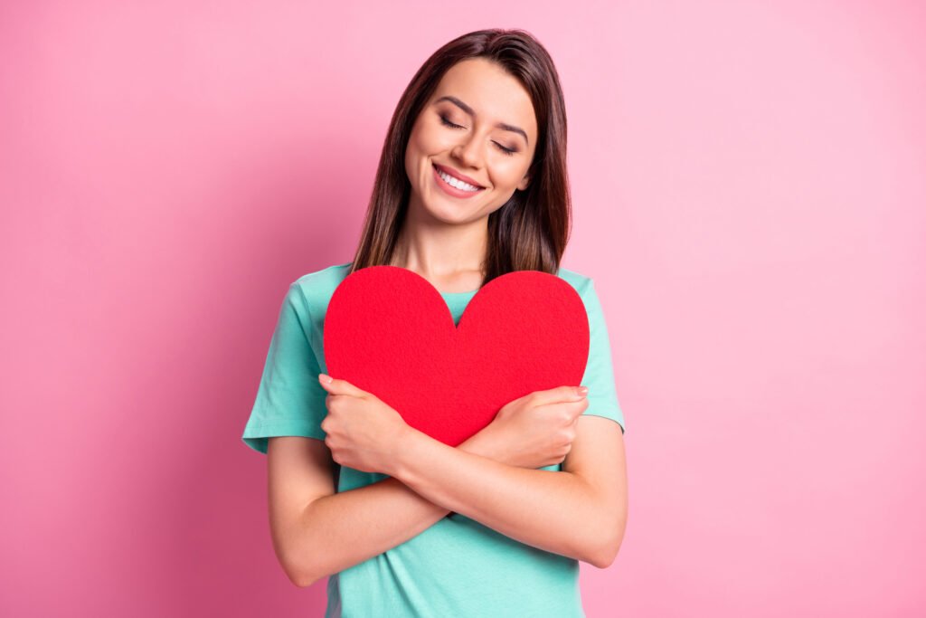 Woman smiling with closed eyes while gently hugging a red heart symbol on a pink background representing self love and emotional healing