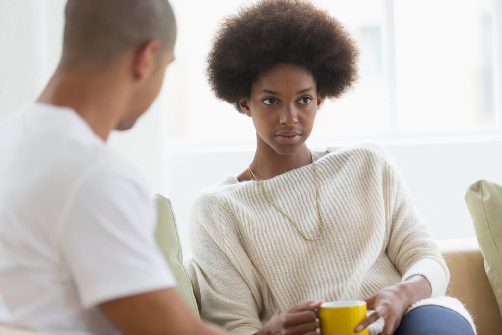 Woman sitting on a couch holding a mug while looking at her partner with a serious and thoughtful expression during a difficult conversation.