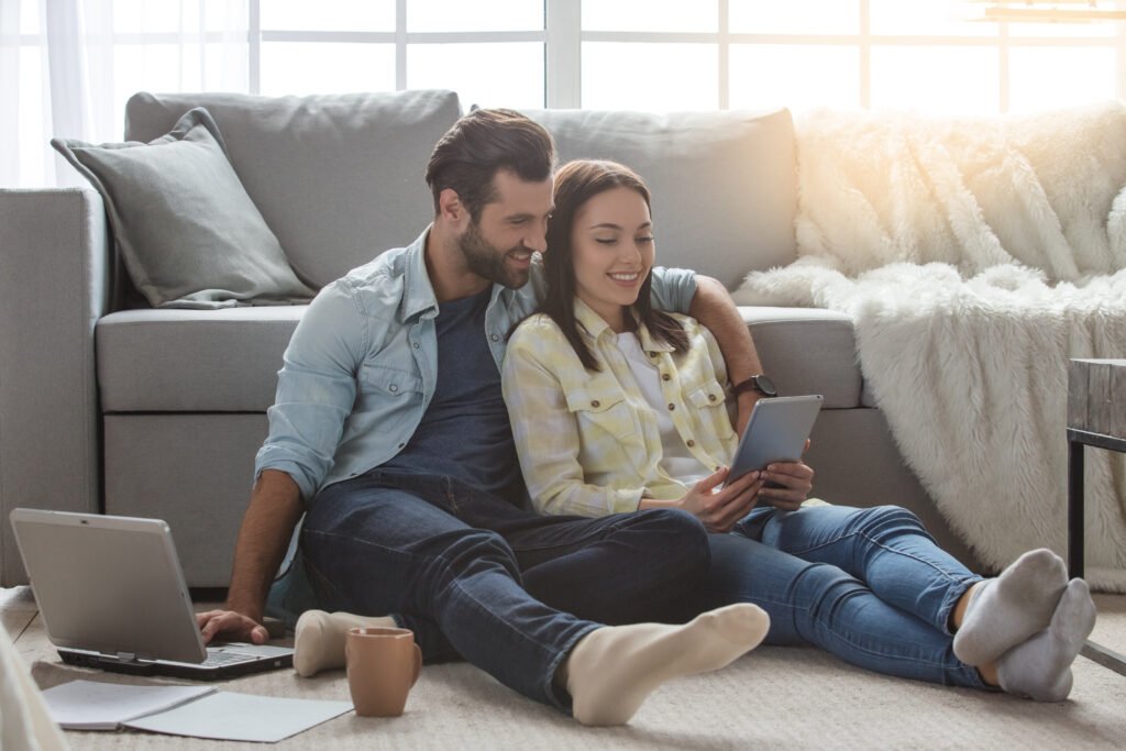 Couple sitting on the floor together at home, smiling and relaxed as they enjoy a quiet moment of connection while looking at a tablet.