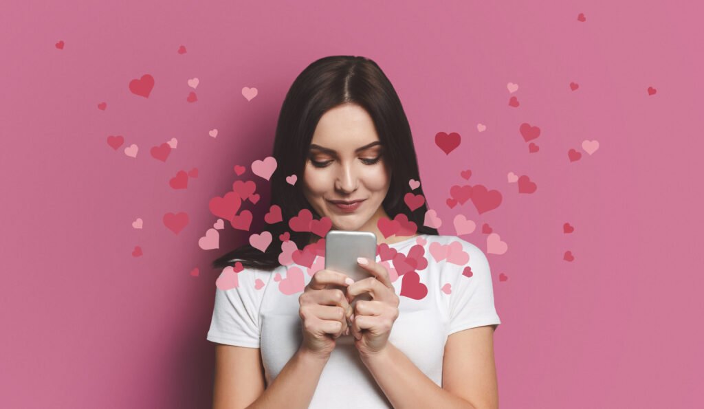 A woman smiling down at her phone with pink heart icons floating around her, symbolizing online dating excitement, romantic messages, and growing connection.