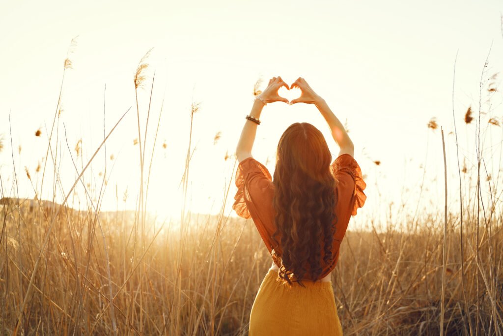 A woman standing in a golden field at sunset forming a heart shape with her hands, symbolizing self love and emotional healing.