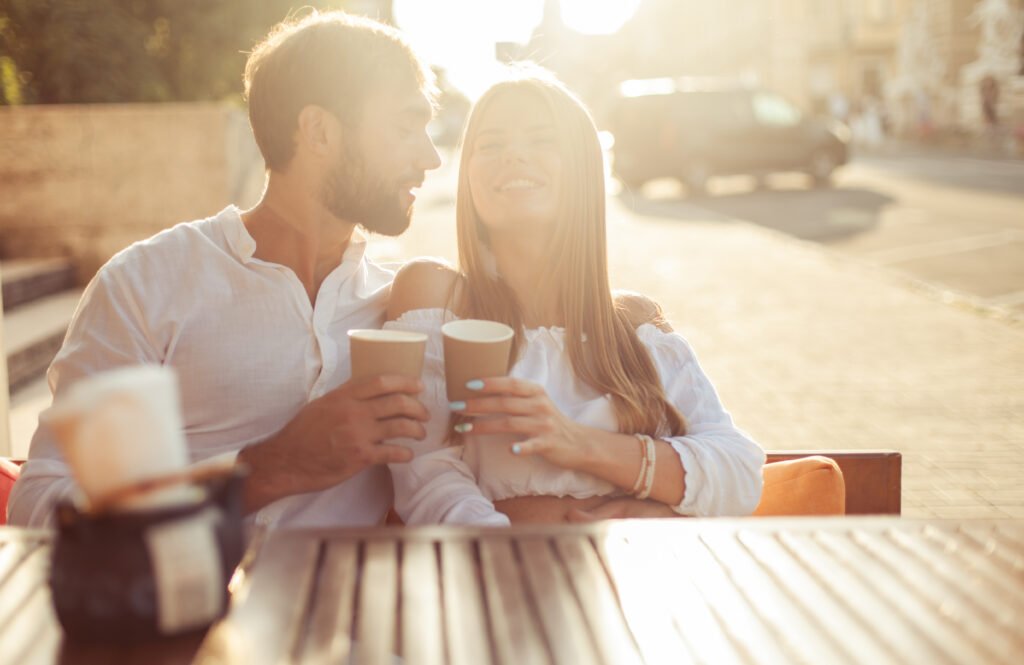 A couple sitting outdoors during sunset, smiling and holding coffee cups while enjoying a warm and relaxed moment together.